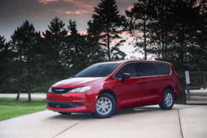 Red 2025 Chrysler Voyager minivan parked on a driveway with trees and a colorful sunset in the background, highlighting its stylish and family-friendly design.
