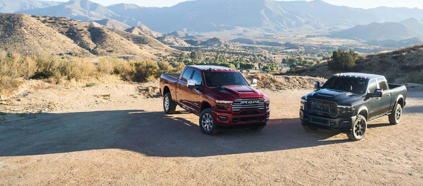 Two RAM 3500 trucks, one red and one black, parked on a scenic dirt overlook with mountains in the background. Available at Bernard Northtown.
