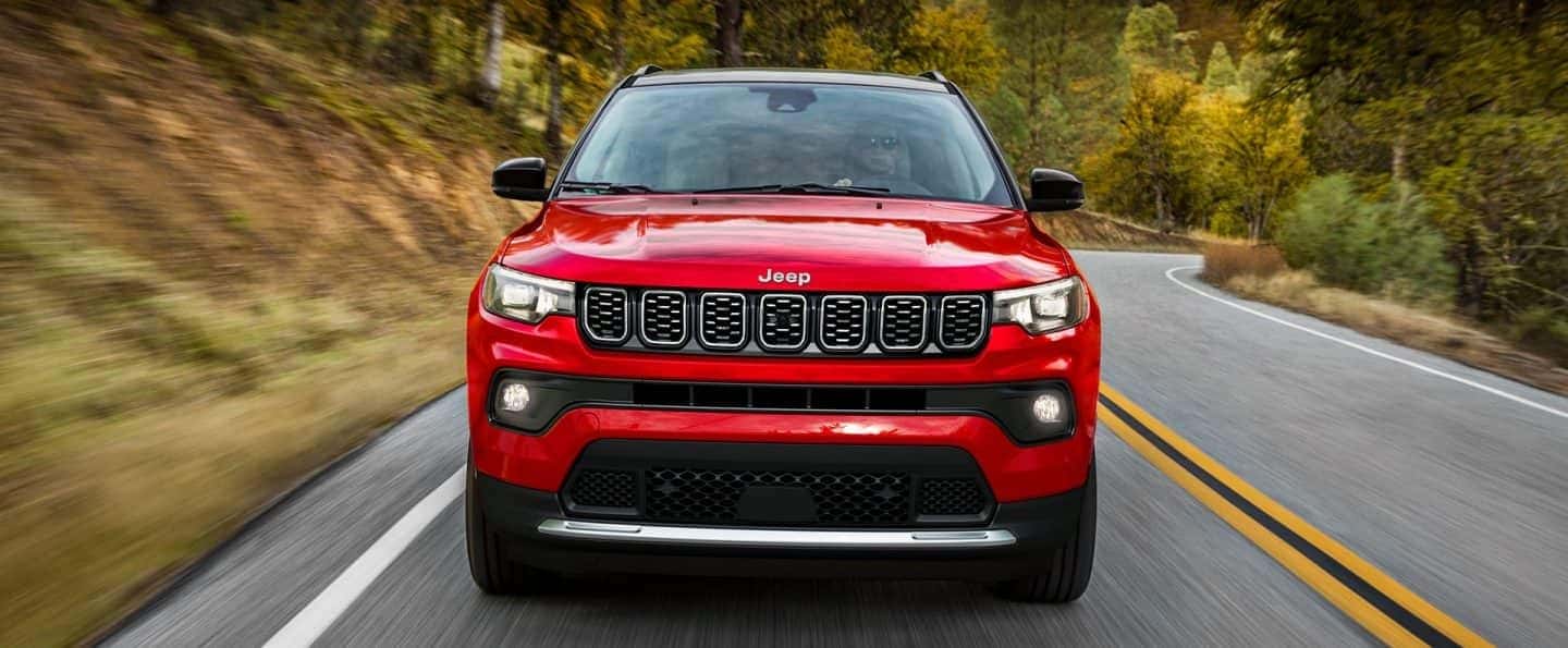 Front view of the red 2025 Jeep Compass driving on a forest-lined road, highlighting its bold grille and rugged performance — test drive today at Bernard's Northtown Jeep in Wisconsin.