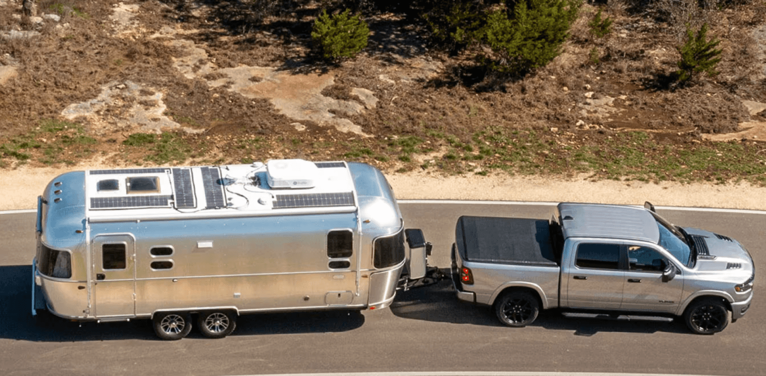 Silver RAM 1500 towing a trailer on a curved road near a wooded area.