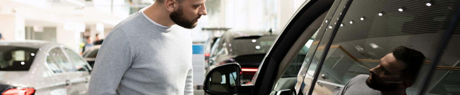 Customer inspecting a vehicle inside a dealership showroom while looking through the side window at the interior features.