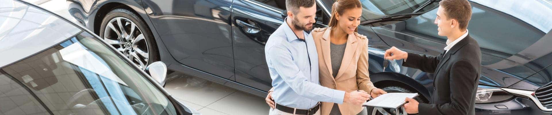 Car salesperson handing over keys to a couple at a dealership while they review paperwork beside a vehicle.