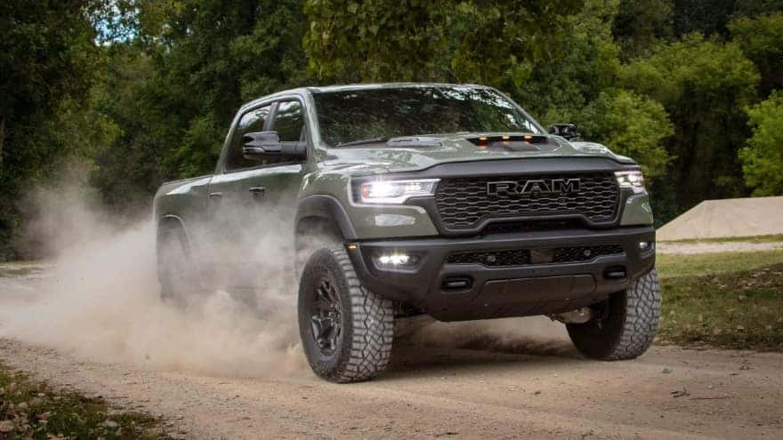 A green RAM 1500 kicking up dust as it drives on a dirt road near a forest.