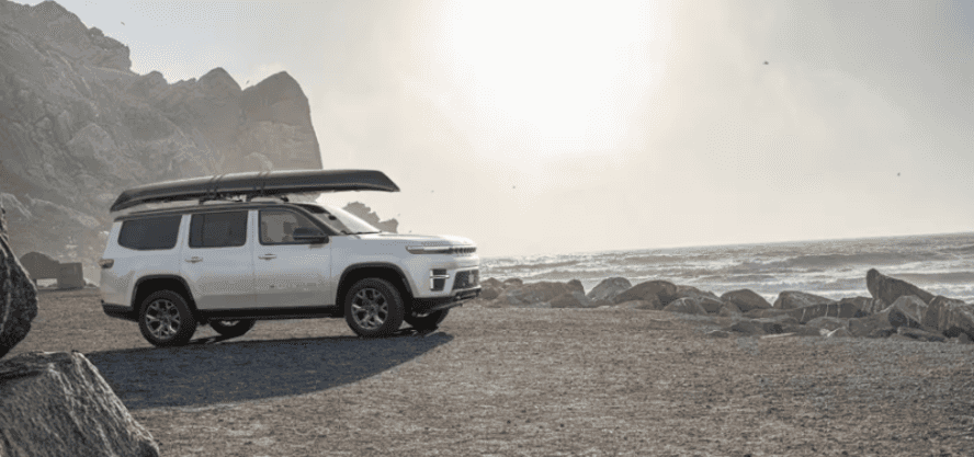 A white Jeep Grand Wagoneer on the beach with a canoe on its roof.
