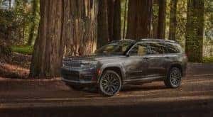 A gray 2021 Jeep Grand Cherokee L is parked beneath large evergreen trees.