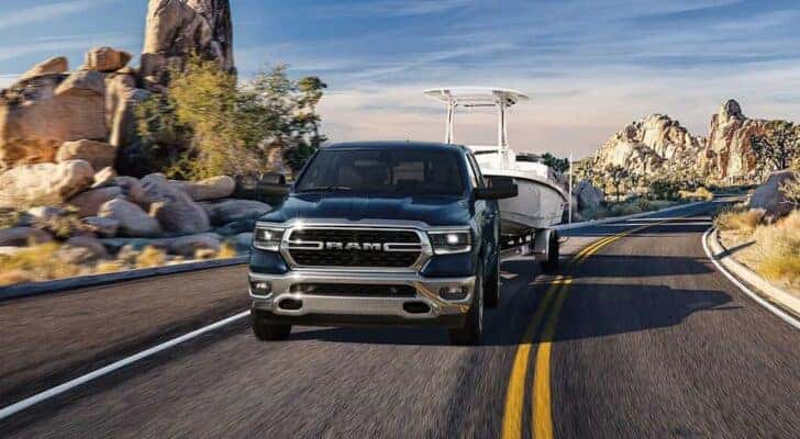 A blue 2023 Ram 1500 is shown towing a boat on a winding road.