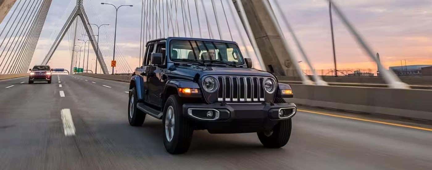 A blue 2023 Jeep Wrangler is shown driving on a bridge.