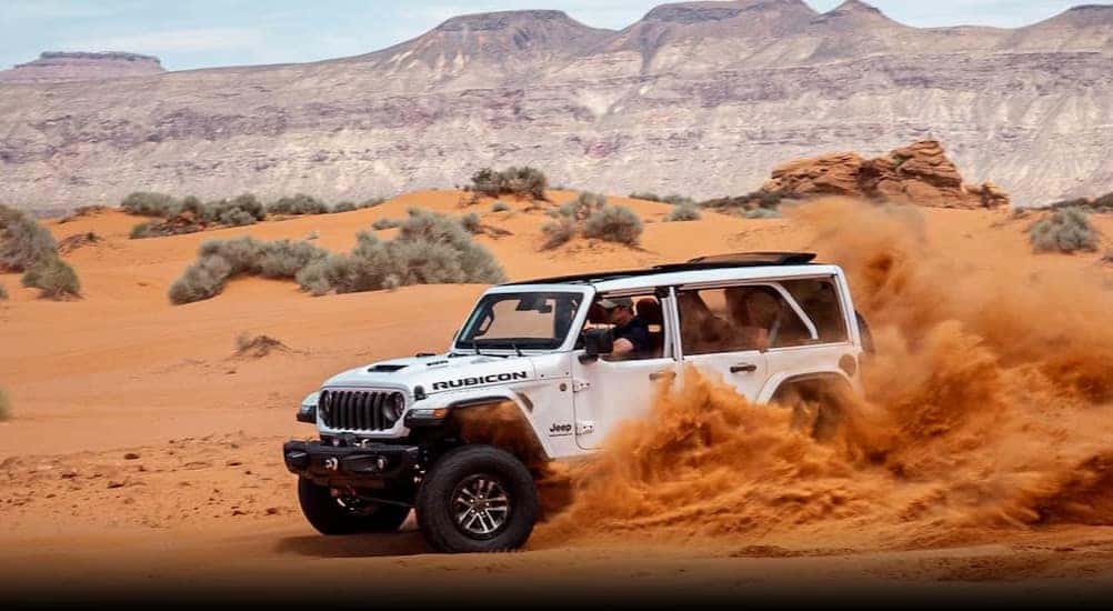A white 2024 Jeep Wrangler Rubicon is shown driving on sand after viewing a Jeep Wrangler for sale near Louisville.
