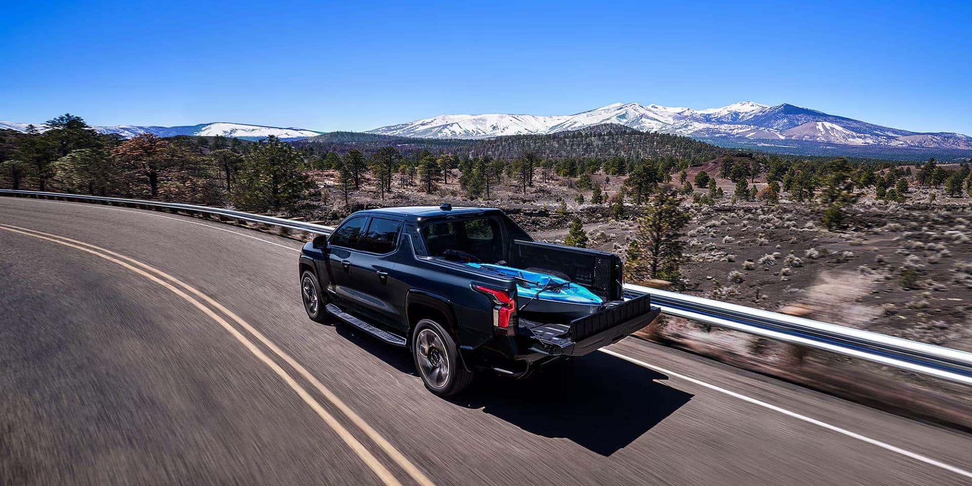 A black Chevrolet Silverado EV driving on a mountain road with outdoor gear in its truck bed.