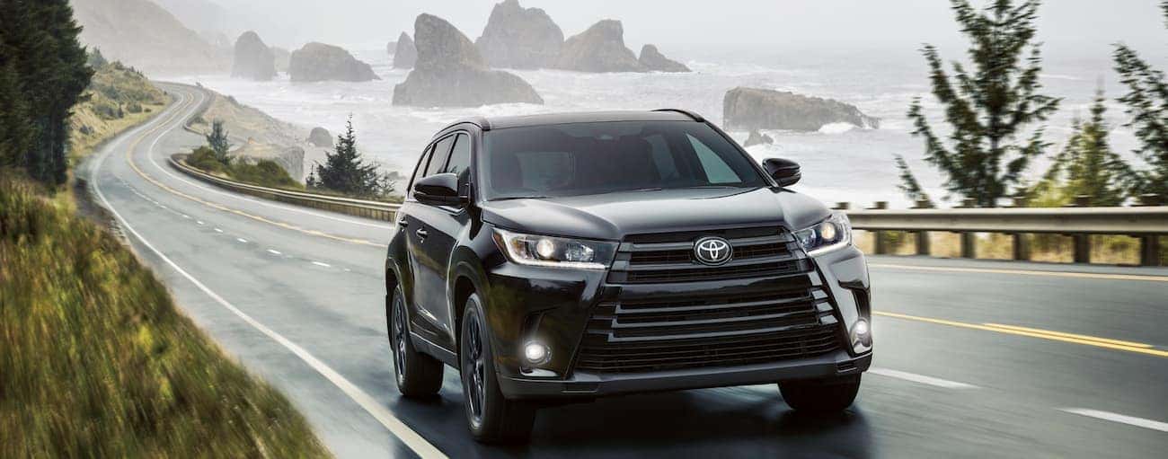 A black 2019 Toyota Highlander drives along a wet road alongside a body of water