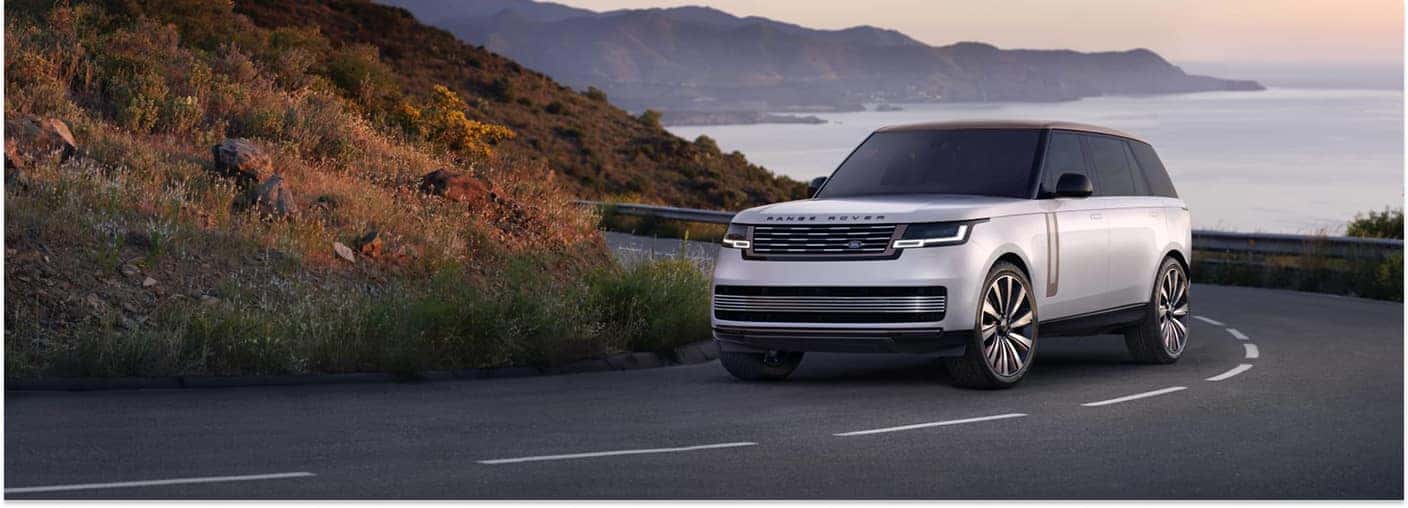 Silver 2025 Range Rover SUV driving on a coastal mountain road in front three-quarter view.