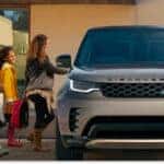 Front view of a silver 2026 Land Rover Discovery parked outside a modern home as a family approaches.