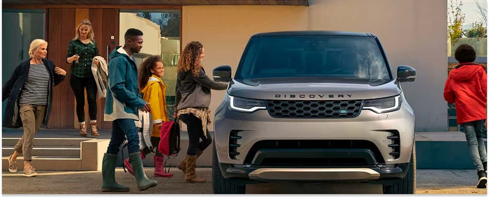 Front view of a silver 2026 Land Rover Discovery parked outside a modern home as a family approaches.