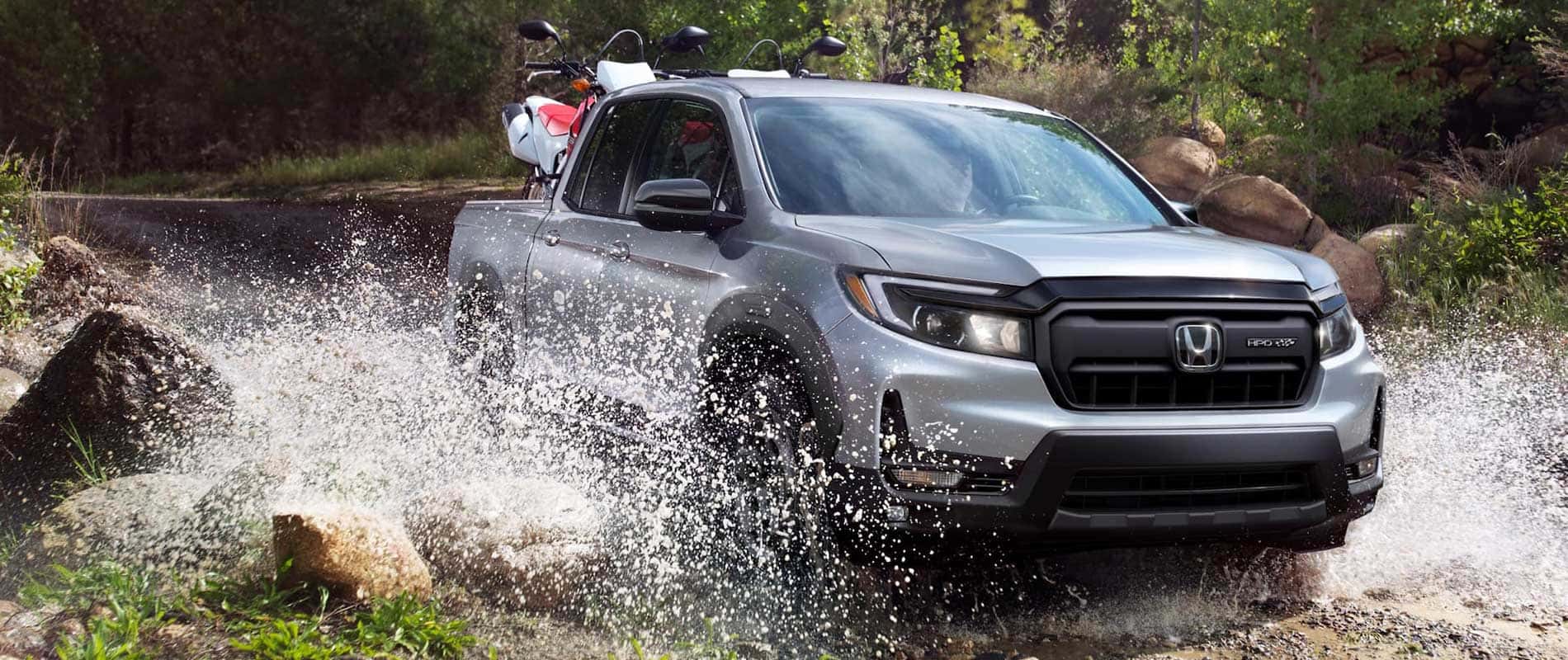 A silver Honda Ridgeline driving through mud while hauling a pair of dirt bikes.