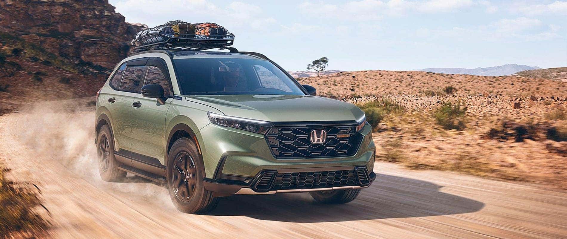 A green Honda CR-V kicking up dust as it drives down a dirt road in a desert.