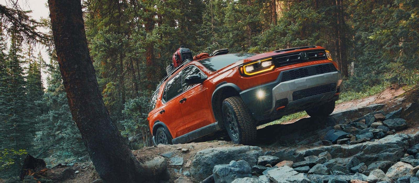 A red-orange Honda Passport driving off-road over rocks in a forest.