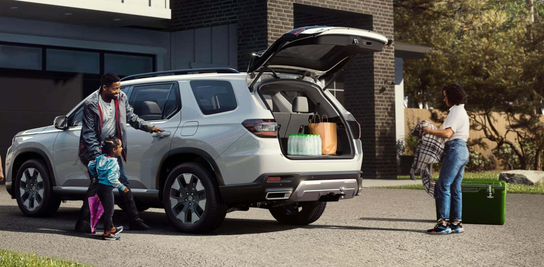 A family of three loading up a silver Honda Pilot in their driveway.