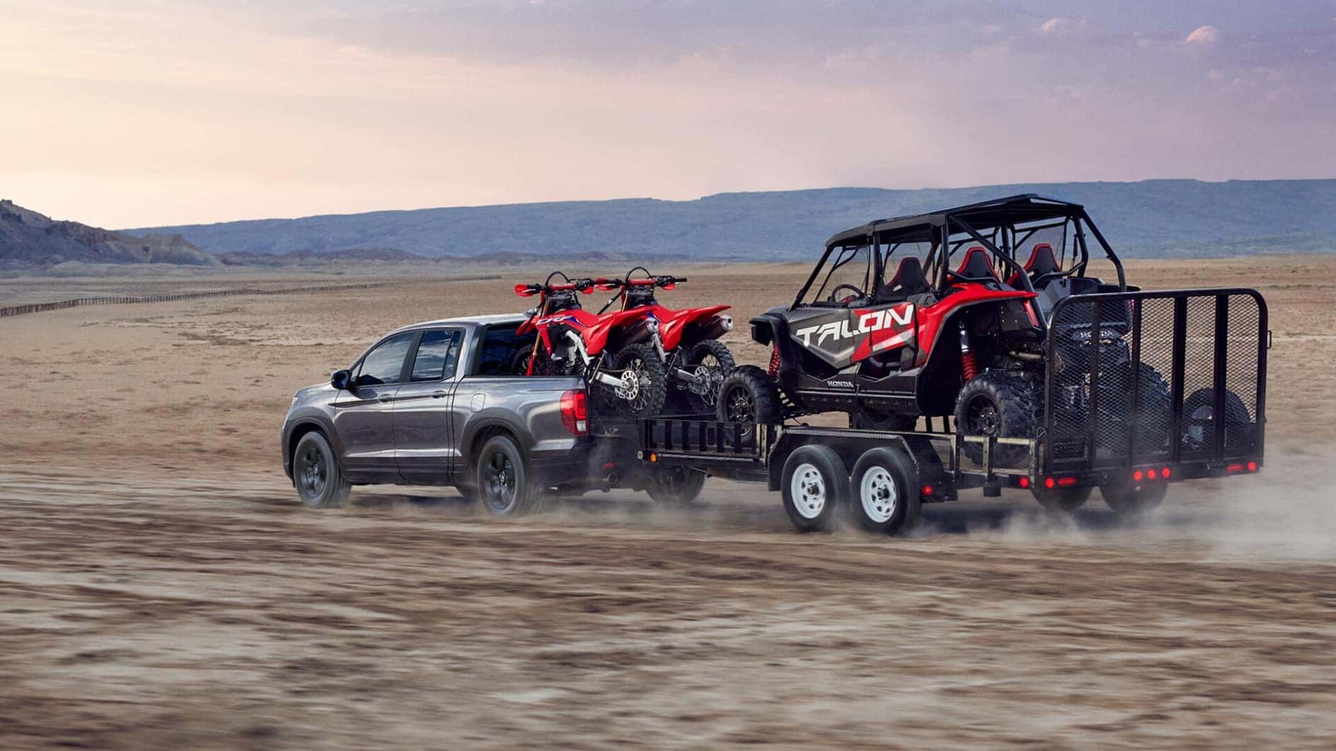 A gray Honda Ridgeline towing a trailer with dirt bikes and a dune buggy off-road through the desert.