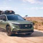 A green Honda CR-V driving on a dirt road through the desert, kicking up dust.