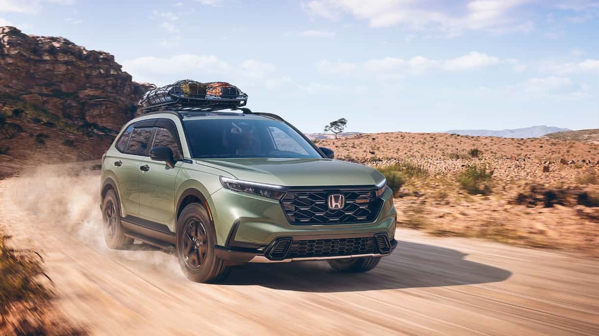 A green Honda CR-V driving on a dirt road through the desert, kicking up dust.