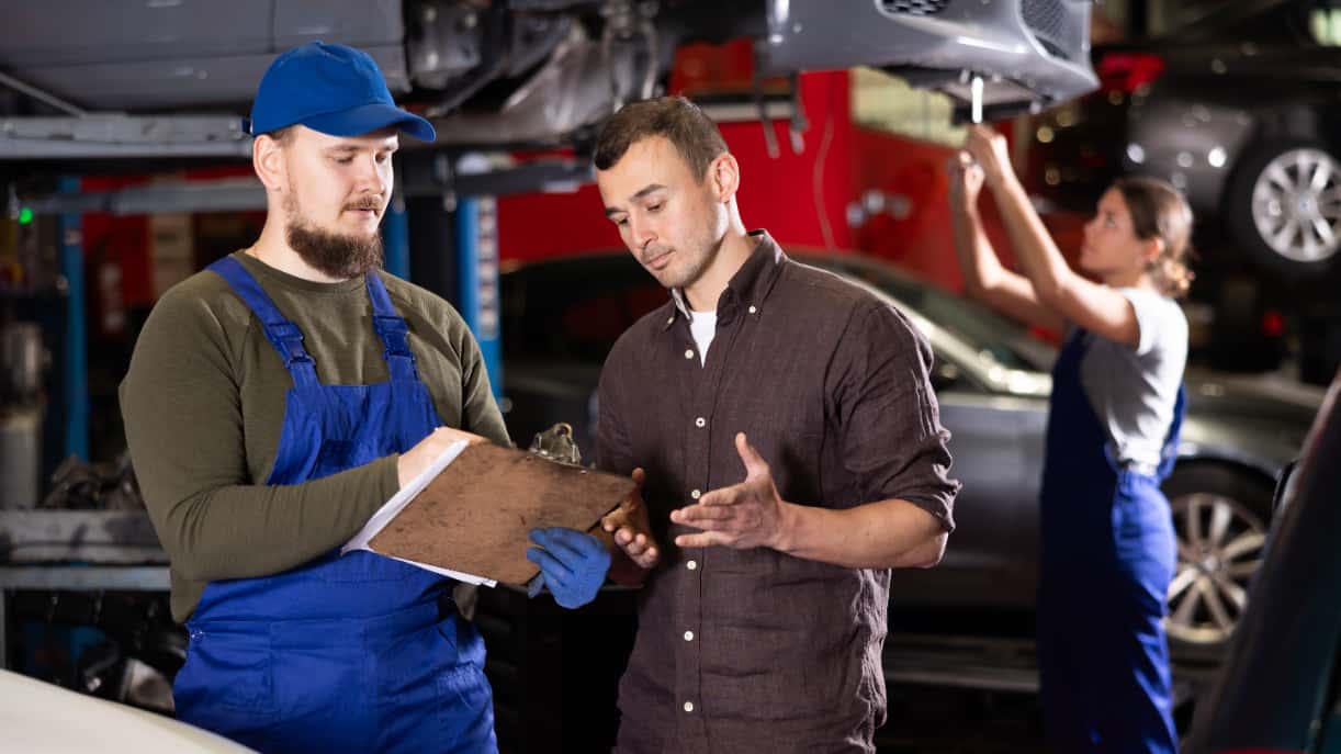 A service technician talking to a customer while another technician works on a car in the background.