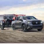 Grey 2026 Honda Ridgeline towing a utility trailer with a UTV on a dirt road in a desert landscape.