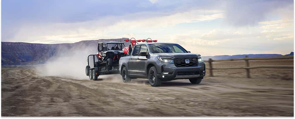 Grey 2026 Honda Ridgeline towing a utility trailer with a UTV on a dirt road in a desert landscape.