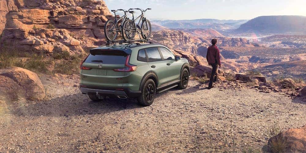 A person standing beside a green 2027 Honda CR-V SUV with a bike rack parked at the edge of a mountain cliff