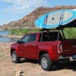 A red 2019 GMC Canyon is parked off-road near a beach with paddle boards on the rack.