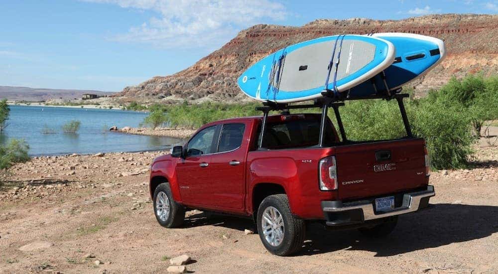 A red 2019 GMC Canyon is parked off-road near a beach with paddle boards on the rack.