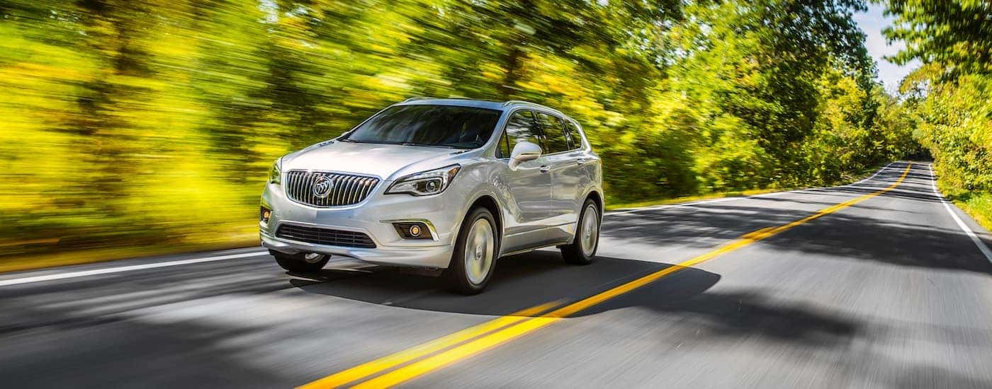 A white 2017 Buick Envision is driving on a sunny, tree-lined road.