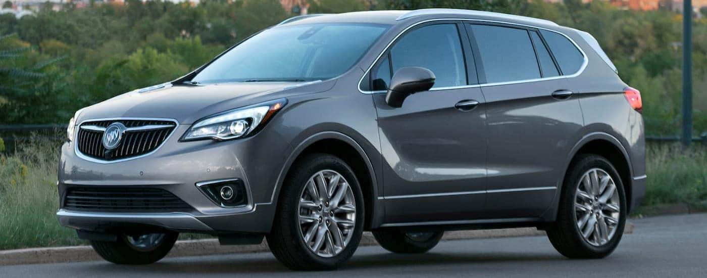 A grey 2020 Buick Envision, popular among the 2020 Buick SUVs, is parked next to trees with a city sky line in the distance. 