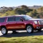 A red 2018 GMC Yukon XL, which is popular among used SUVs for sale, is driving on a sunny road past trees.