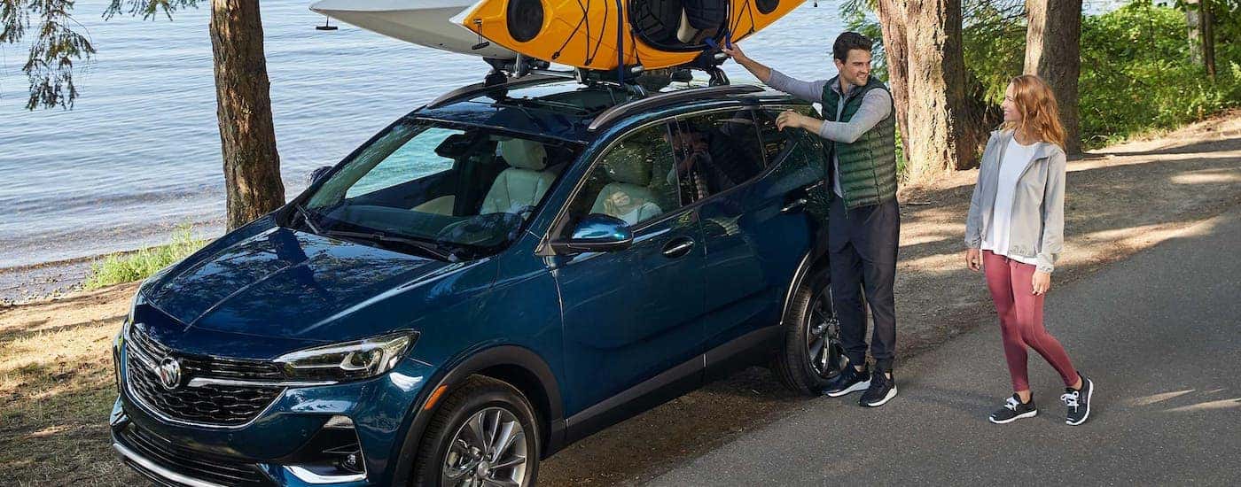 A couple is loading kayaks on top of a dark blue 2020 Buick Encore GX which is parked in front of water.