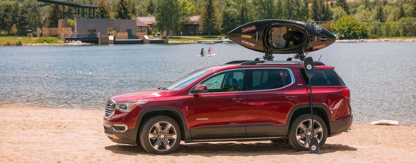 A red 2018 GMC Acadia with a kayak on the roof is parked in front of a lake and mountains.