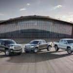 A silver 2015 used GMC Canyon, a grey 2014 used GMC Sierra 1500, and a grey 2015 used GMC Sierra Denali 3500 HD are parked in front of a glass faced building.