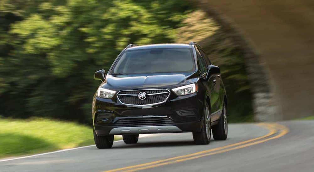 A black 2021 Buick Encore is driving on a tree-lined road past an arch bridge.
