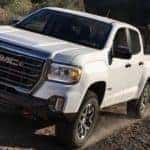 A white 2022 GMC Canyon AT4 is shown pulling a trailer with garden equipment on a dirt road.