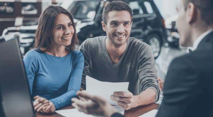 A couple is shown talking to a salesperson at a car dealership.