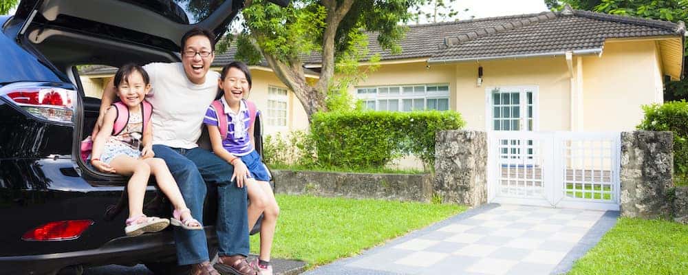 Parent with children seated on the back of their car, in front of a house with bright green foliage