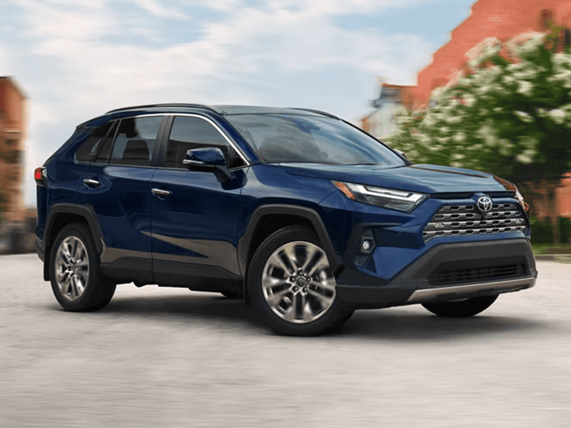 A blue Toyota RAV4 is parked in an urban setting on a concrete road. Trees and buildings are behind the vehicle.