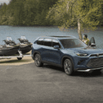 A man stands next to the driver's side door of a Toyota Grand Highlander Hybrid. The vehicle is blue and it's parked on concrete. A lake is in the background, and a speedboat is shown hitched to the vehicle, as if it is about to be backed down into the water.