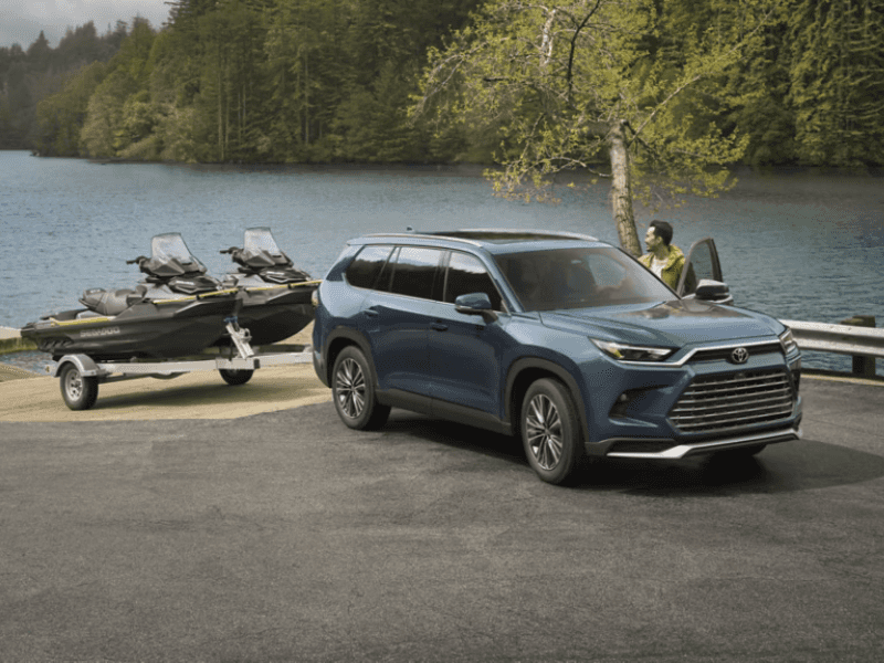 A man stands next to the driver's side door of a Toyota Grand Highlander Hybrid. The vehicle is blue and it's parked on concrete. A lake is in the background, and a speedboat is shown hitched to the vehicle, as if it is about to be backed down into the water.