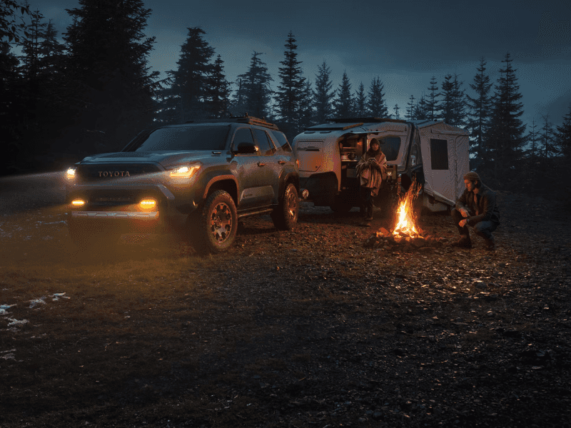 A green Toyota 4Runner sits at a campsite next to a campfire. It's twilight and the headlights of the vehicle are on. A man sits next to the fire, and a woman stands next to a small camper that is hitched to the rear of the 4Runner.