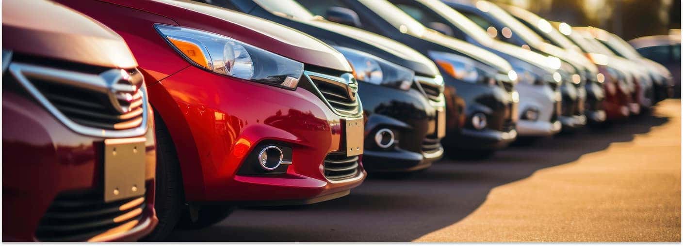 > Close-up front view of a long row of various multi-colored cars parked on an outdoor lot, with a bright red vehicle in sharp focus in the foreground under warm sunlight.