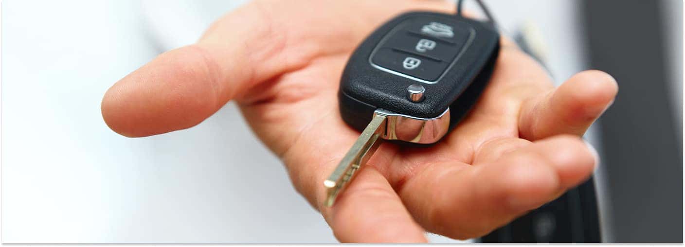 > Close-up view of a person's open hand holding a black car key fob with the metal key blade extended against a bright, blurred background.