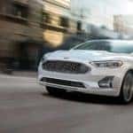 A white 2019 Ford Fusion is driving down a city block near Carbondale, IL.